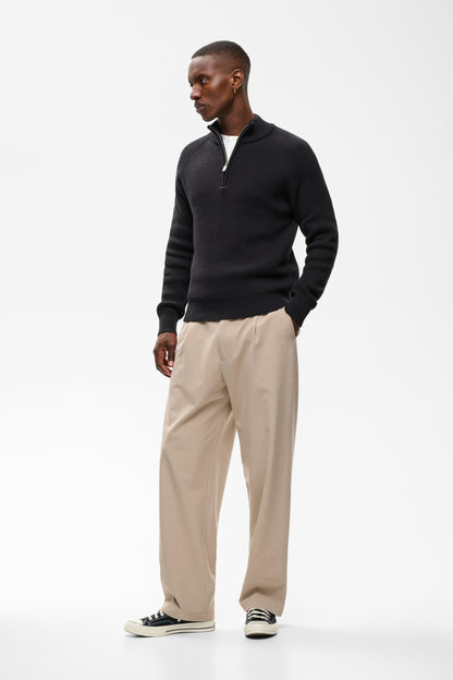 A man modeling a black long-sleeve quarter-zip pullover sweater, paired with beige trousers and black-and-white sneakers, against a plain white studio background.