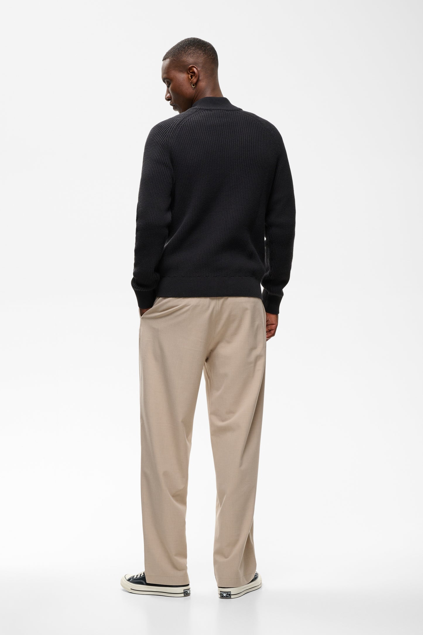 A man viewed from behind wearing a black long-sleeve knit sweater, beige trousers, and black-and-white sneakers, standing against a clean white studio background.