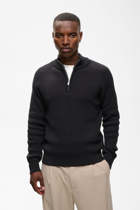 A man wearing a black ribbed knit quarter-zip sweater with a stand collar, styled with beige trousers, against a plain white studio background.