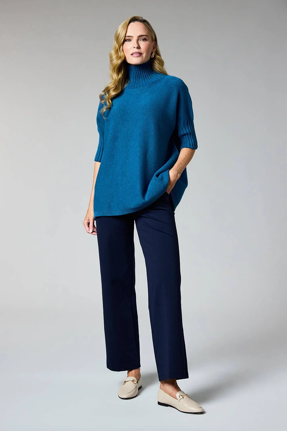 A woman modeling a teal blue mock-neck knit sweater with elbow-length sleeves, paired with dark navy wide-leg trousers and beige loafers, against a light gray studio background.