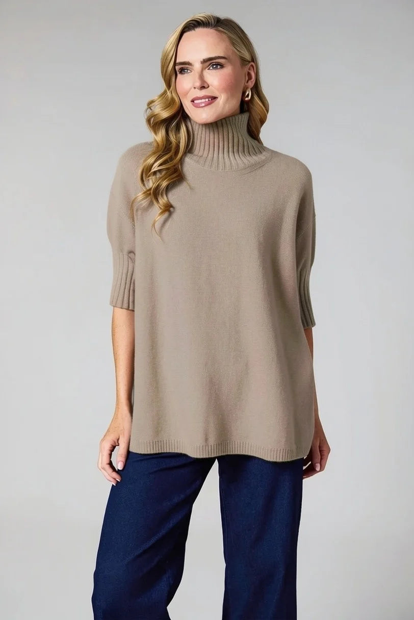 A woman modeling a taupe knit turtleneck sweater with elbow-length sleeves and dark navy wide-leg pants, photographed against a neutral light gray studio background.