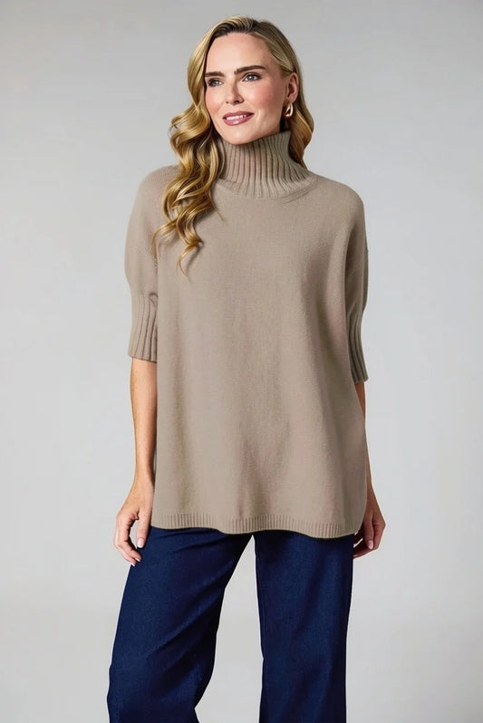 A woman modeling a taupe knit turtleneck sweater with elbow-length sleeves and dark navy wide-leg pants, photographed against a neutral light gray studio background.