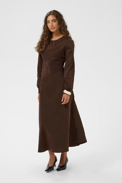 A woman modeling a long-sleeved brown midi dress with a smocked bodice and a contrasting white cuff, paired with black pointed-toe heels, against a light gray studio background.