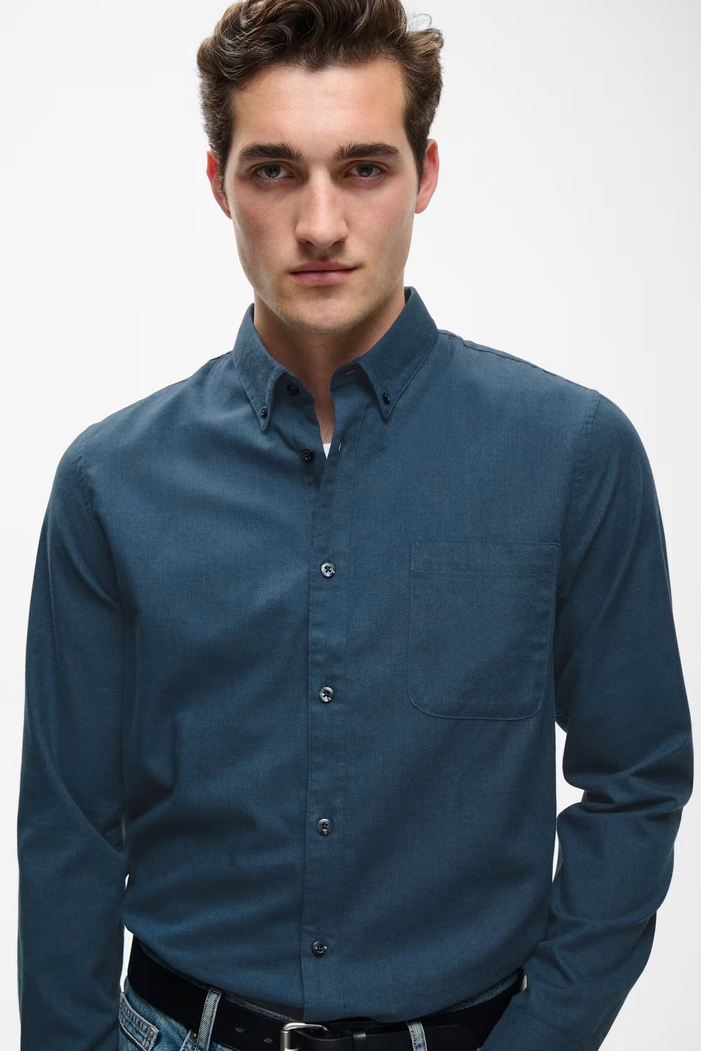 A man wearing a dark blue long-sleeve button-down shirt, paired with a black belt and blue jeans, photographed against a light gray studio background.