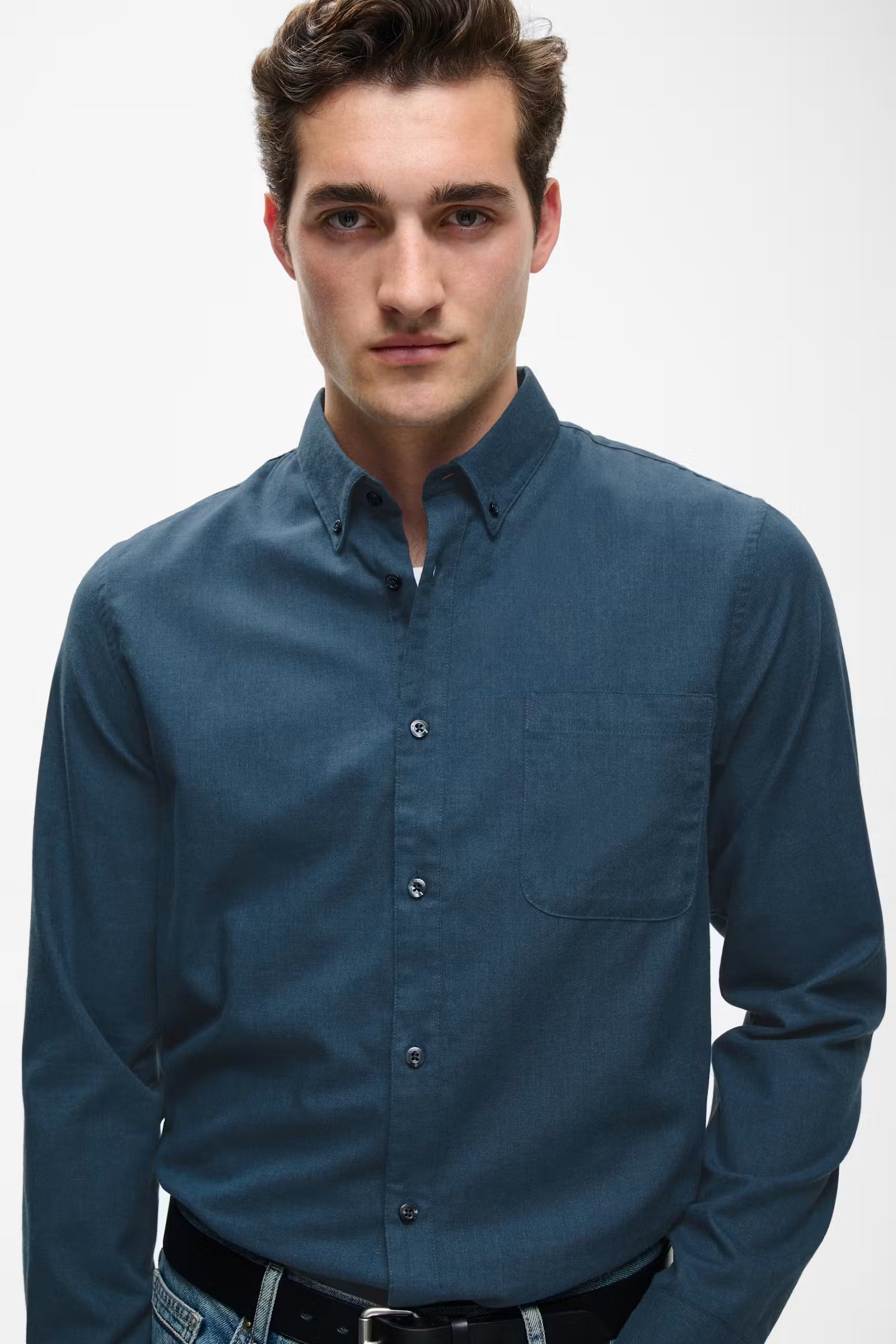 A man wearing a dark blue long-sleeve button-down shirt, paired with a black belt and blue jeans, photographed against a light gray studio background.