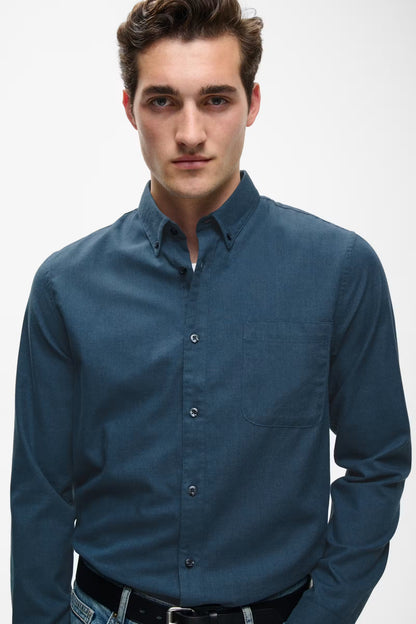 A man wearing a dark blue long-sleeve button-down shirt, paired with a black belt and blue jeans, photographed against a light gray studio background.