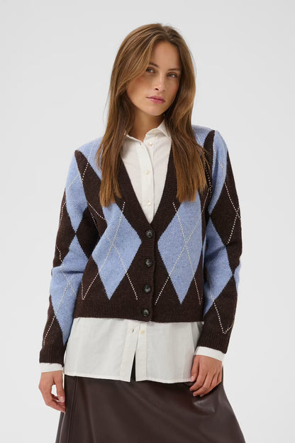 A woman wearing a brown and light blue argyle knit cardigan over a white collared shirt and dark brown skirt, photographed against a light gray studio background.