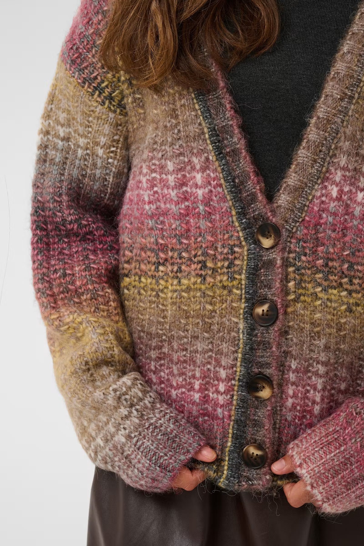 A close-up of a person wearing a chunky knit cardigan with a multicolor plaid pattern in dusty pink, beige, brown, and muted yellow, featuring dark brown buttons and a dark gray top underneath.