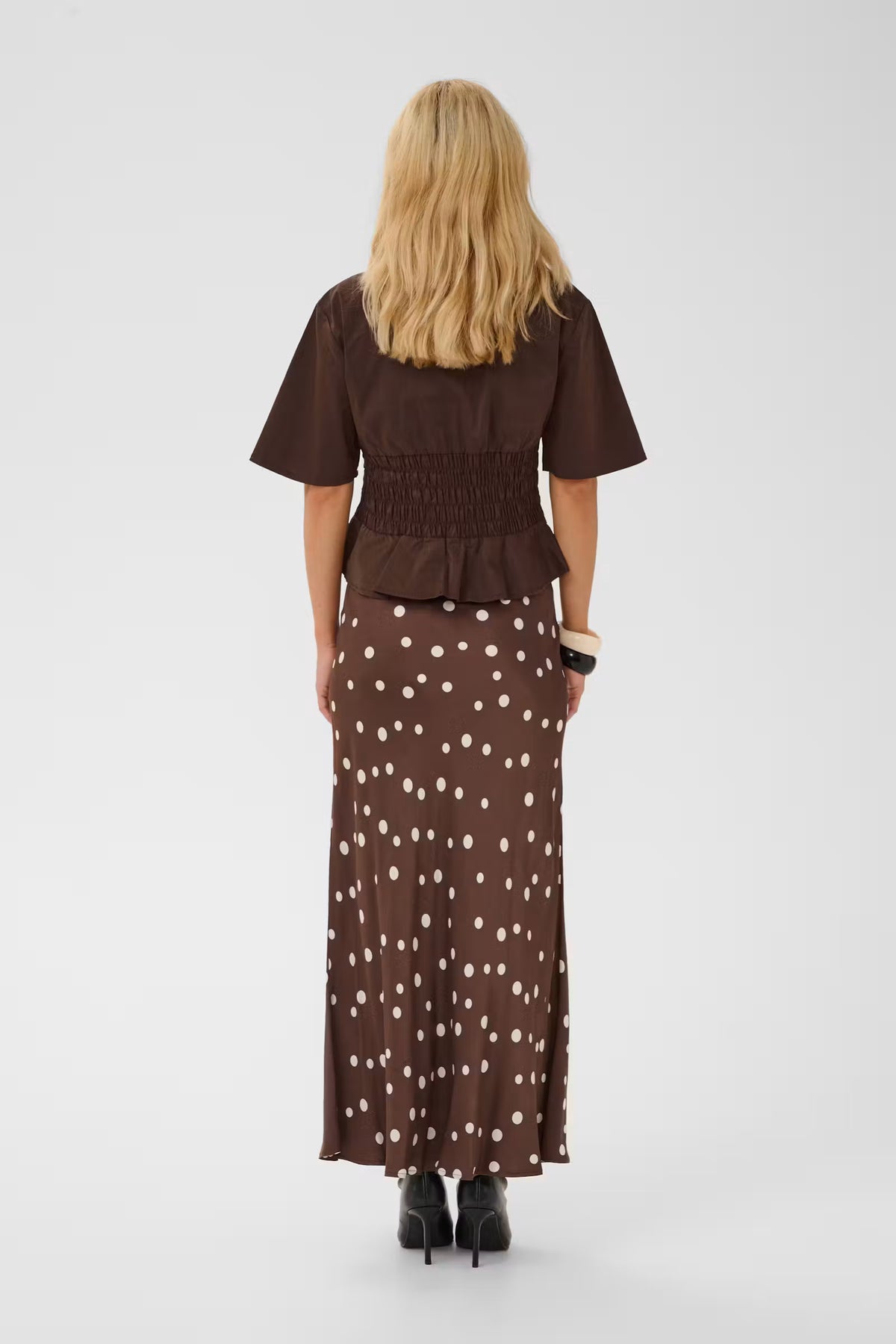 A woman viewed from the back wearing a brown short-sleeve smocked top and a matching brown maxi skirt with white polka dots, standing against a light gray studio background.