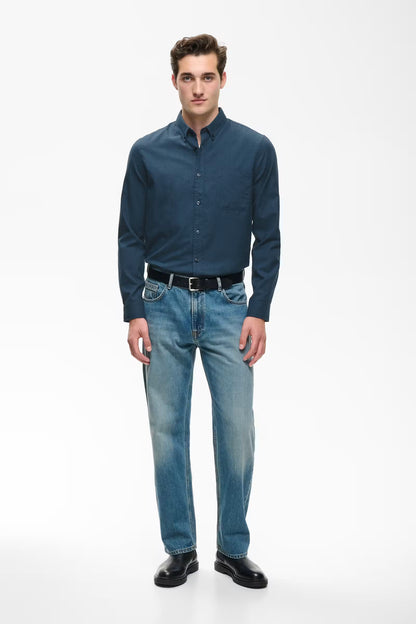 A man modeling a dark blue long-sleeve button-down shirt and light blue jeans with a black belt and black shoes, standing against a plain white studio background.