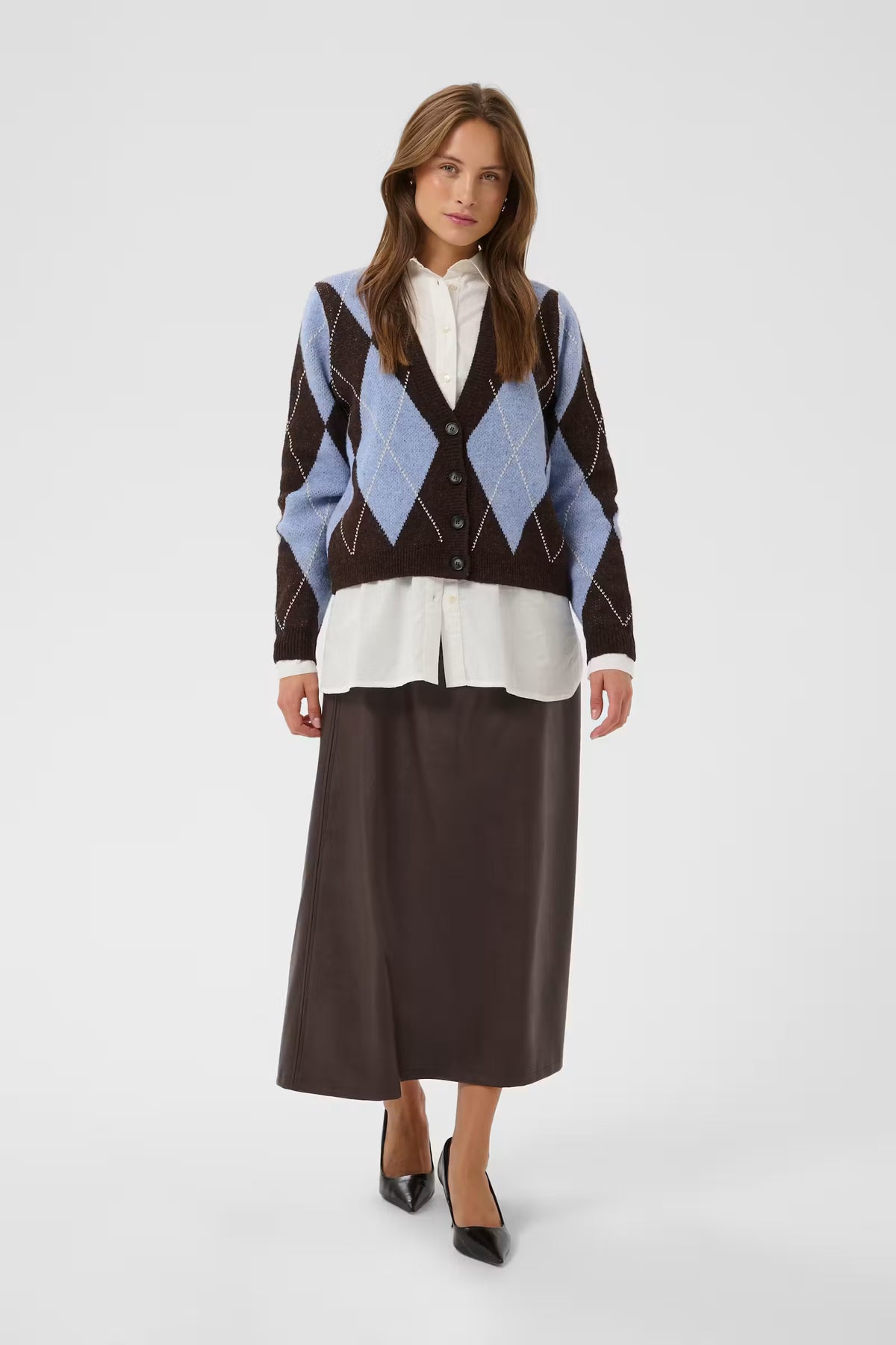 A woman models a brown and light blue argyle cardigan over a white collared shirt and a dark brown midi skirt, paired with black pointed-toe heels, against a light gray studio background.