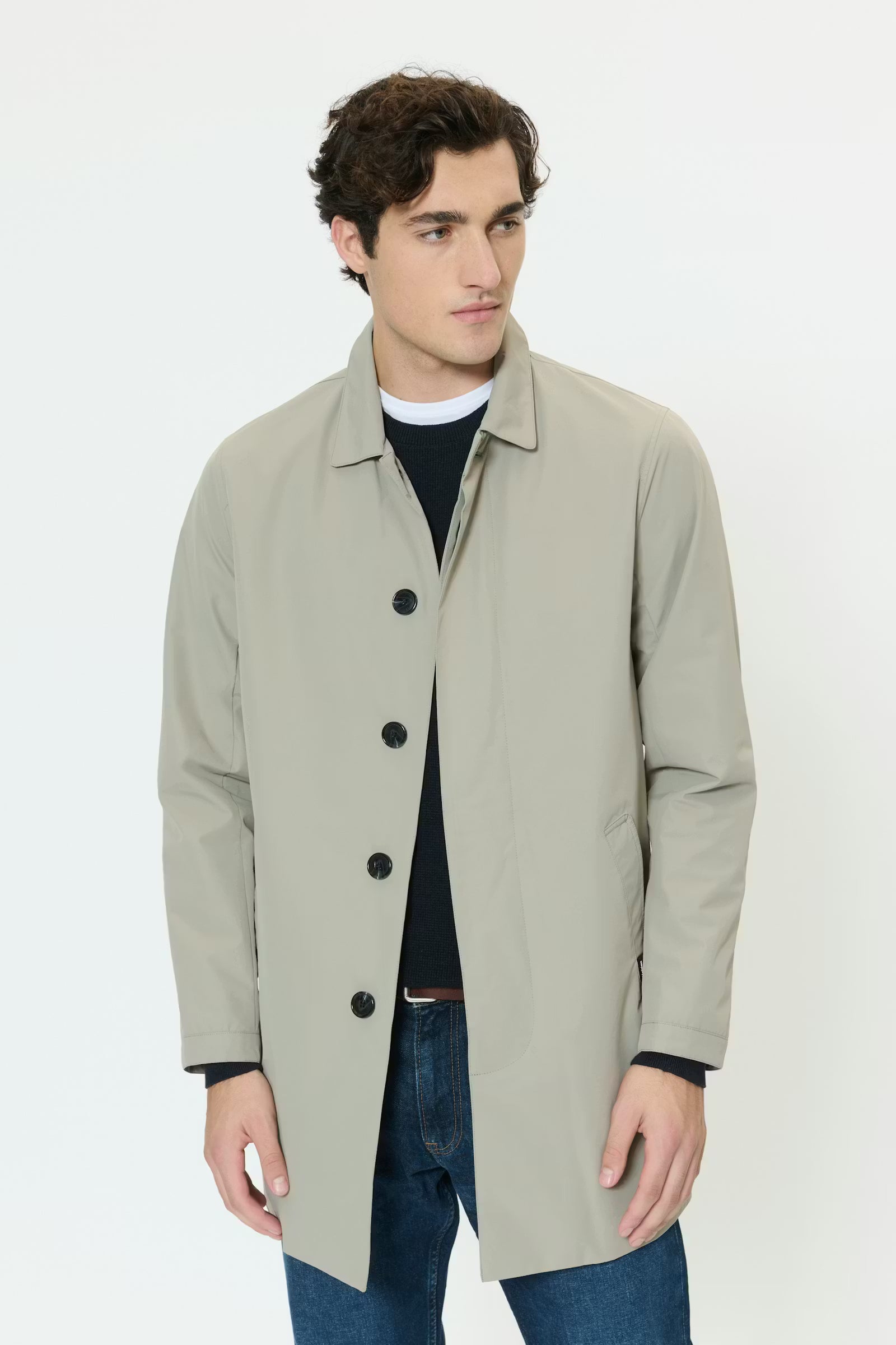 Man wearing a light gray coat over a black shirt and blue jeans on a white background