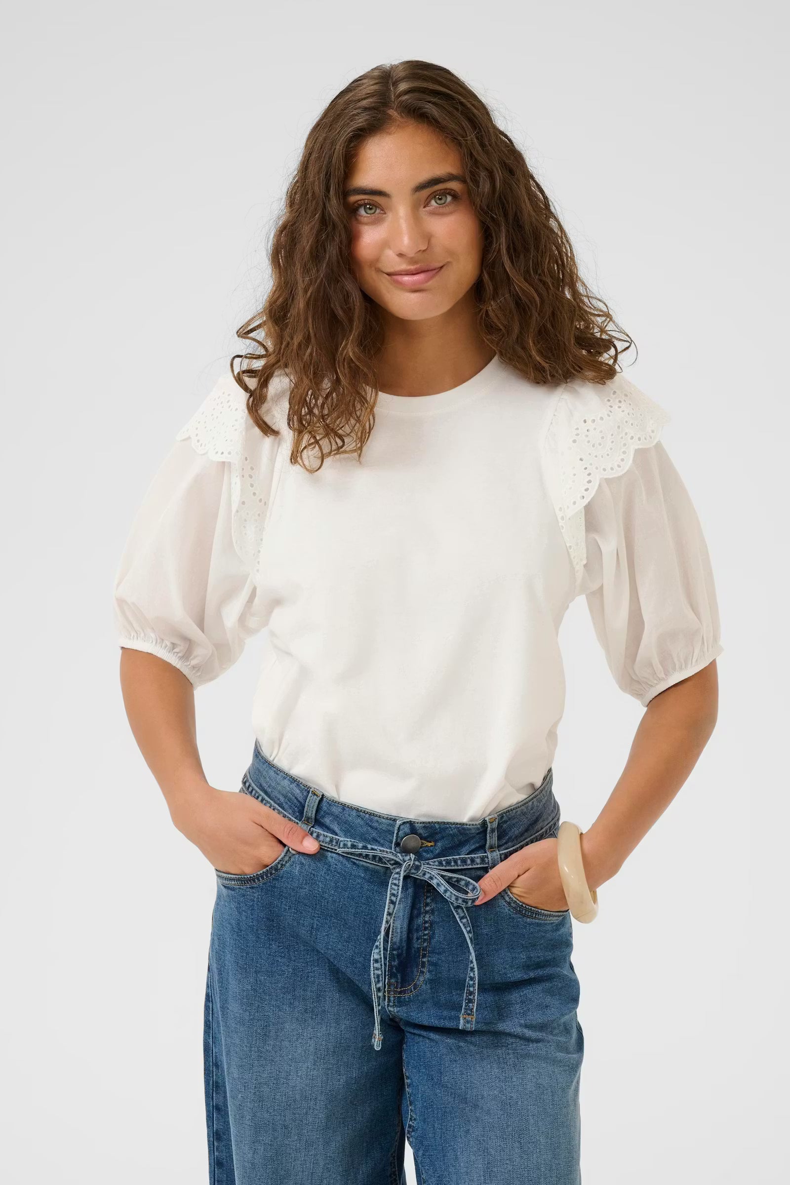 Woman wearing a white blouse with lace details and blue jeans on a plain background