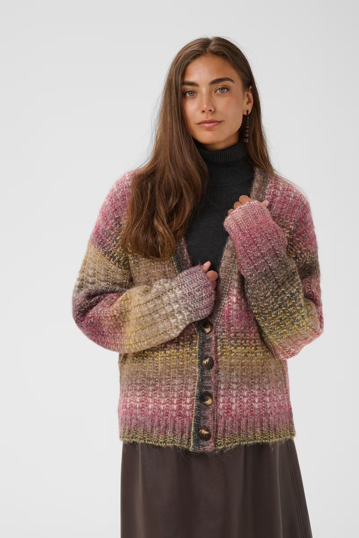 A woman modeling a chunky knit cardigan with a variegated color pattern of pink, beige, and olive green, worn over a black turtleneck and dark skirt against a light gray studio background.