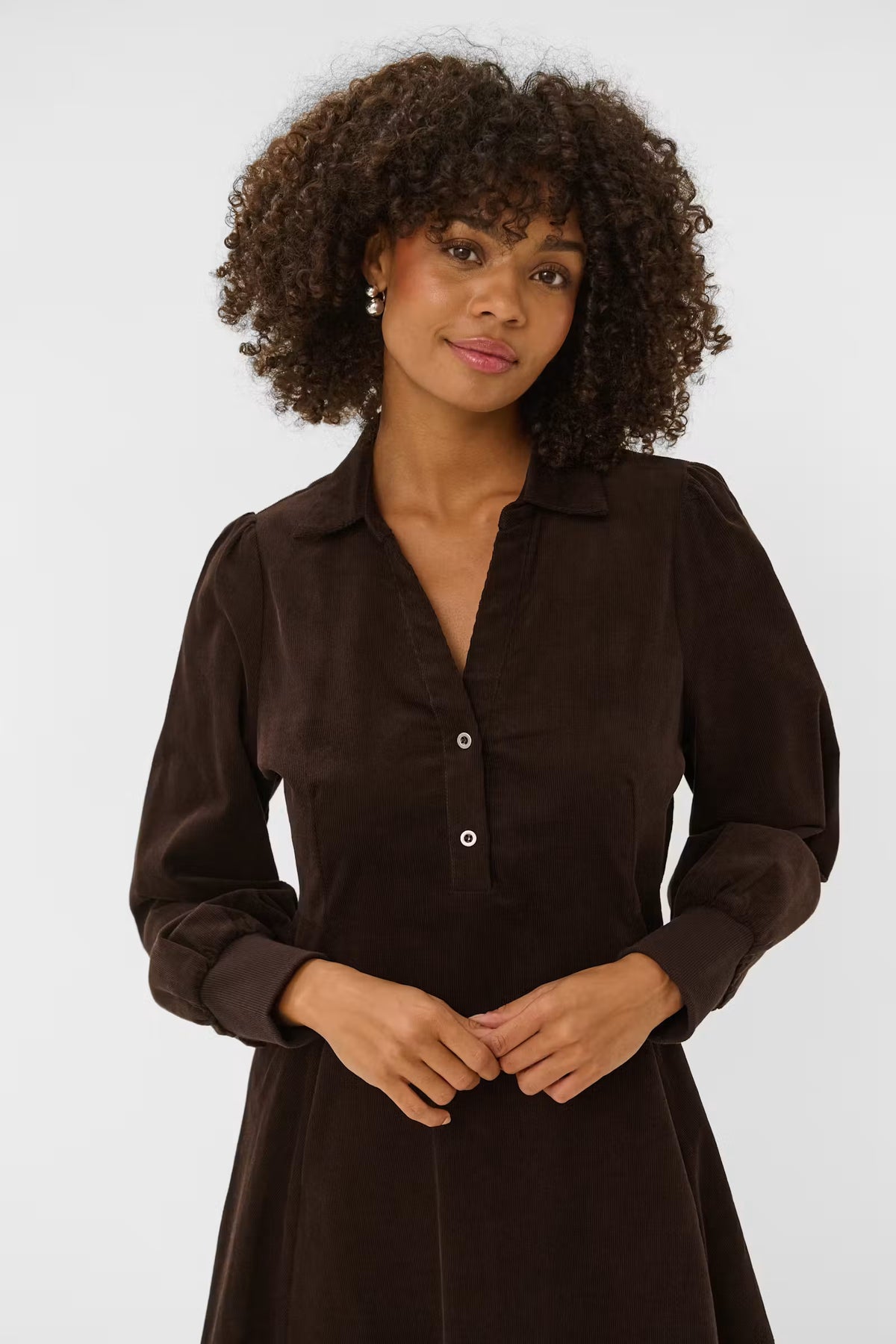 A woman with curly dark hair wearing a dark brown long-sleeve collared shirt dress with button details, standing against a light gray studio background.