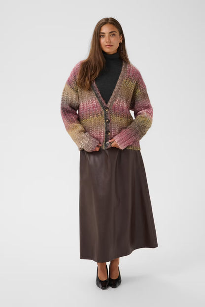 A woman stands against a light gray studio background wearing a multicolor knit cardigan with pink, purple, yellow, and green tones over a black turtleneck, paired with a dark brown midi skirt and black heels.