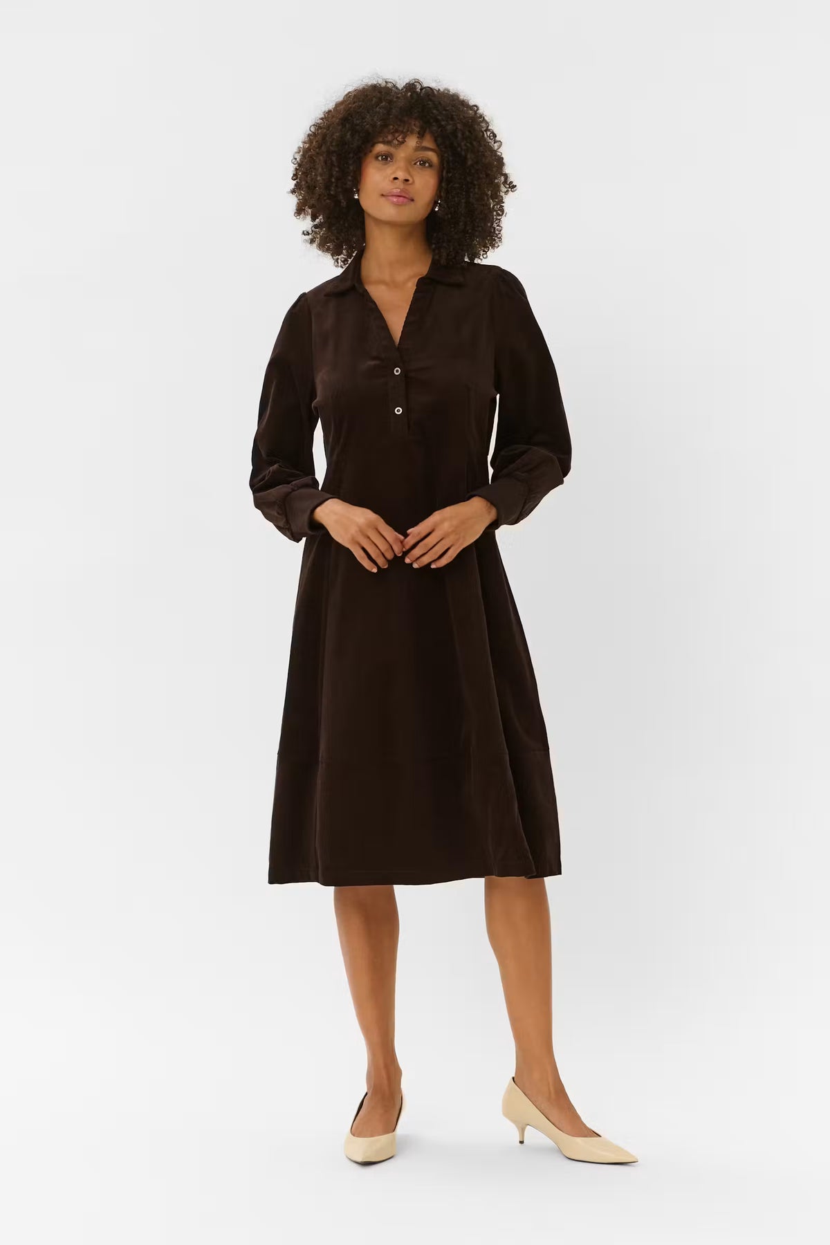 A woman with curly dark hair models a dark brown long-sleeve midi dress with a buttoned V-neckline, standing against a plain light gray studio background.