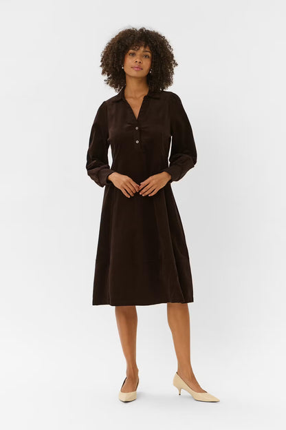 A woman with curly dark hair models a dark brown long-sleeve midi dress with a buttoned V-neckline, standing against a plain light gray studio background.