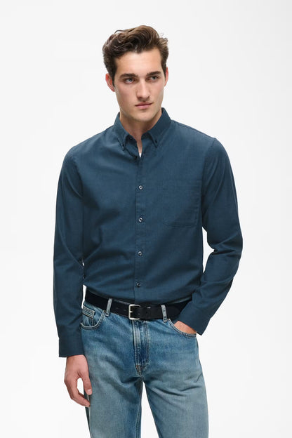 A man wearing a dark blue long-sleeve button-down shirt and blue jeans, standing against a plain white studio background.
