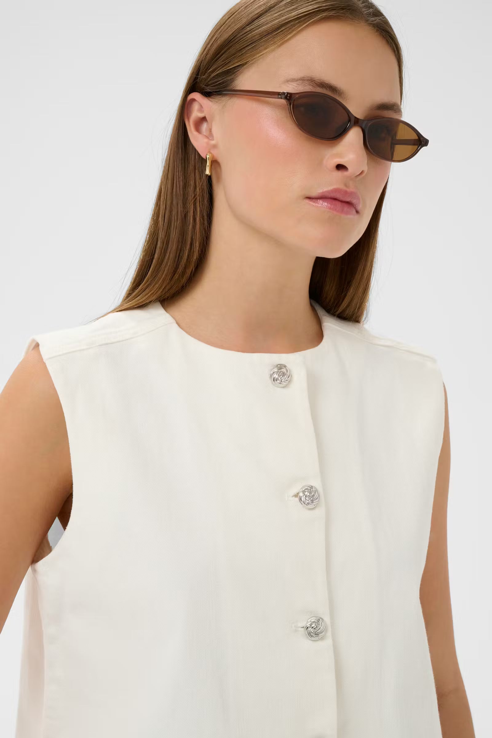 Woman wearing a white sleeveless top with floral buttons against a plain background