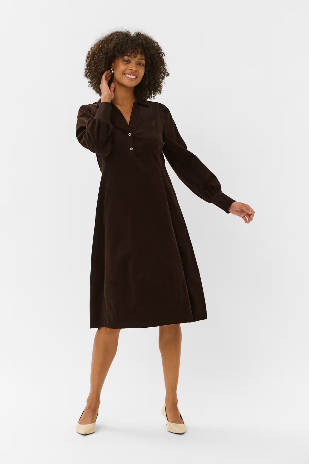 A woman modeling a dark brown long-sleeve midi dress with a buttoned V-neckline, photographed against a plain light gray studio background.