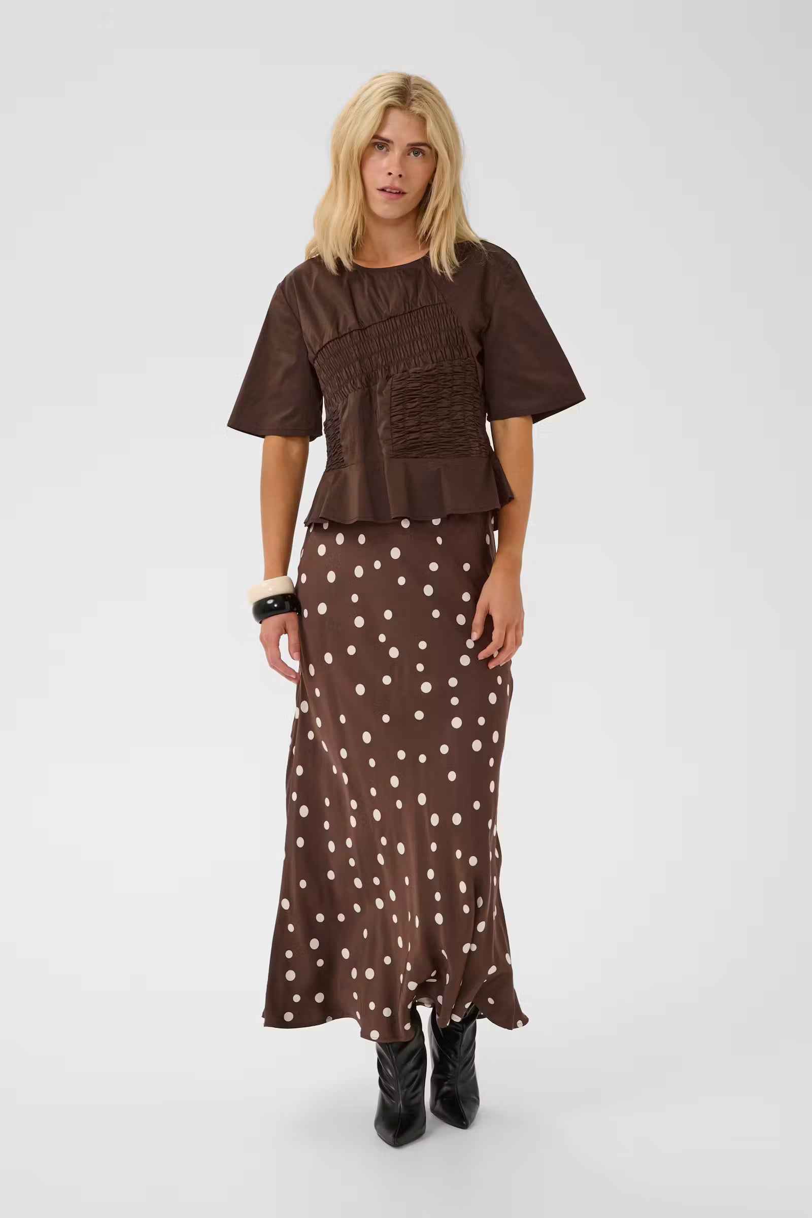 A blonde model wearing a brown short-sleeve top with a smocked bodice and a matching brown maxi skirt with white polka dots, paired with black ankle boots, against a light gray studio background.