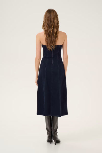 A woman viewed from behind wearing a strapless dark navy midi dress and black knee-high boots against a plain light studio background.