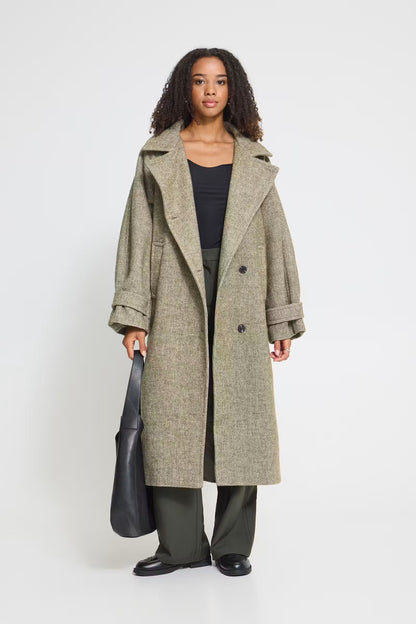 A model stands against a light gray studio background wearing a long, double-breasted olive-gray coat over a black top and dark green wide-leg trousers, while holding a black leather tote bag.