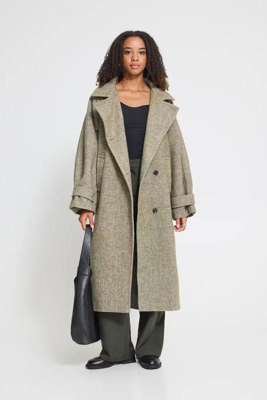 A model stands against a light gray studio background wearing a long, double-breasted olive-gray coat over a black top and dark green wide-leg trousers, while holding a black leather tote bag.