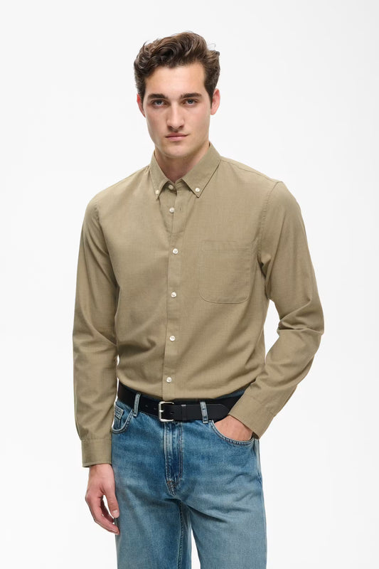 A man wearing a light brown long-sleeve button-down shirt with a chest pocket, paired with blue jeans and a black belt, standing against a plain white studio background.