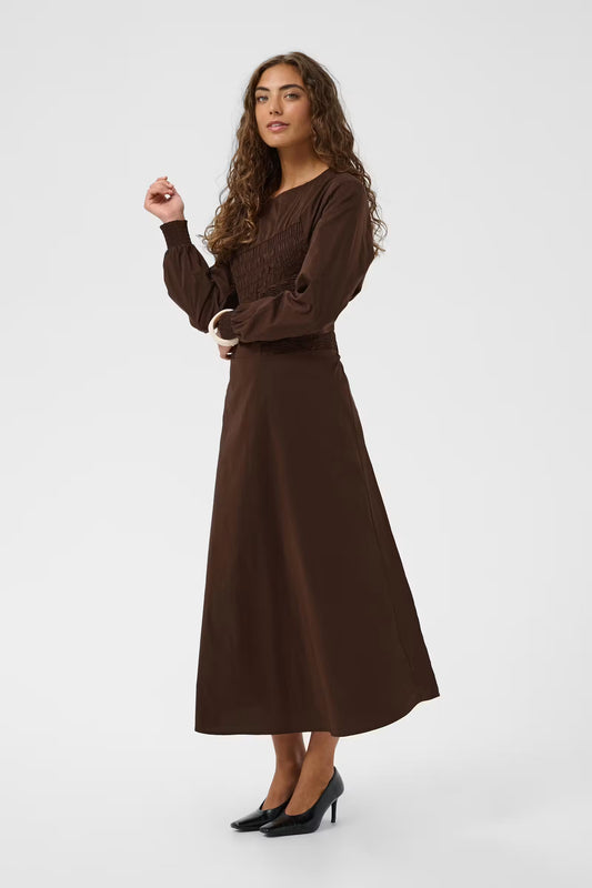 A woman modeling a long-sleeved brown midi dress with a gathered waist, paired with black heels, against a plain light gray studio background.