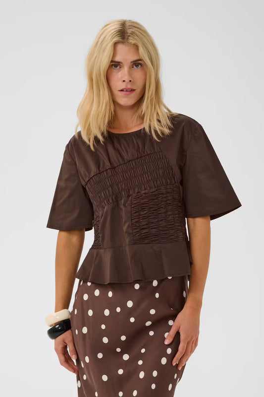 A blonde model wearing a dark brown short-sleeve top with a smocked bodice and ruffle hem, paired with a matching brown polka-dot skirt, against a light gray studio background.