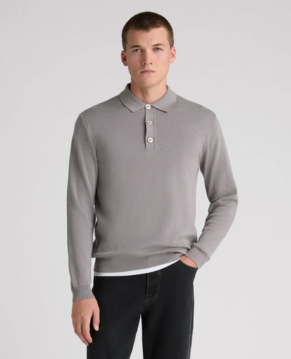 A man wearing a light gray long-sleeve polo sweater and dark jeans, standing against a plain light gray studio background.