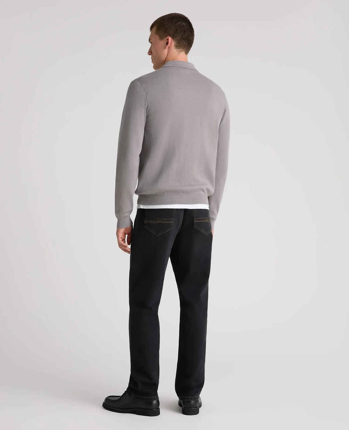 A man shown from the back wearing a light gray long-sleeve knit sweater, dark charcoal jeans, and black shoes, photographed against a clean light gray studio background.