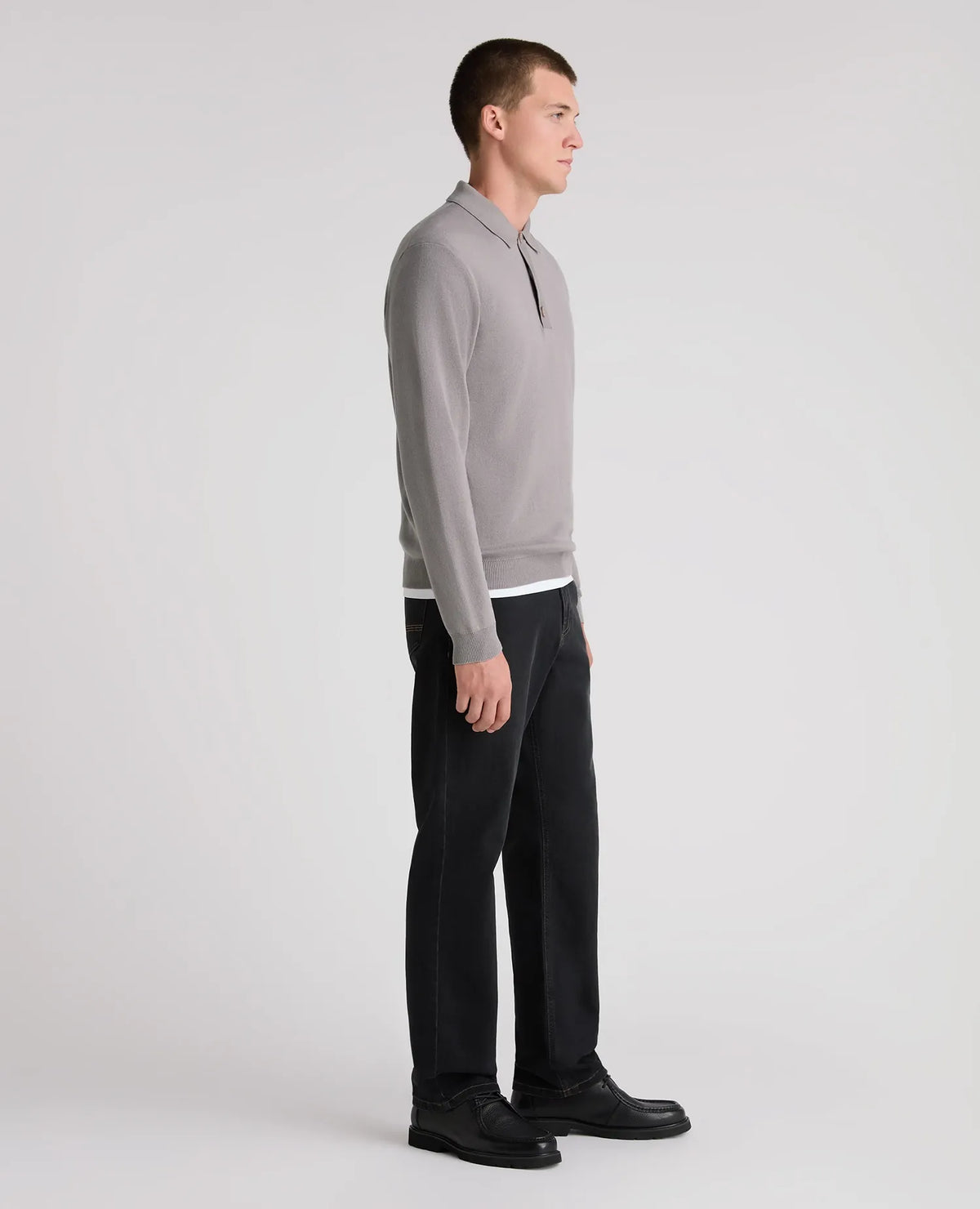A man in a light gray long-sleeve quarter-zip polo sweater, black trousers, and black shoes, shown in profile against a plain light gray studio background.