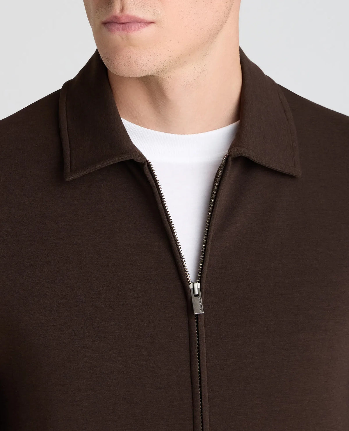 Close-up of a man wearing a dark brown zip-up collar jacket over a white crew-neck t-shirt, against a light gray background.
