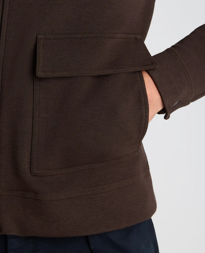 Close-up of a dark brown fleece jacket with a large front flap pocket and ribbed hem, worn by a model whose hand is in the pocket, against a light gray background.