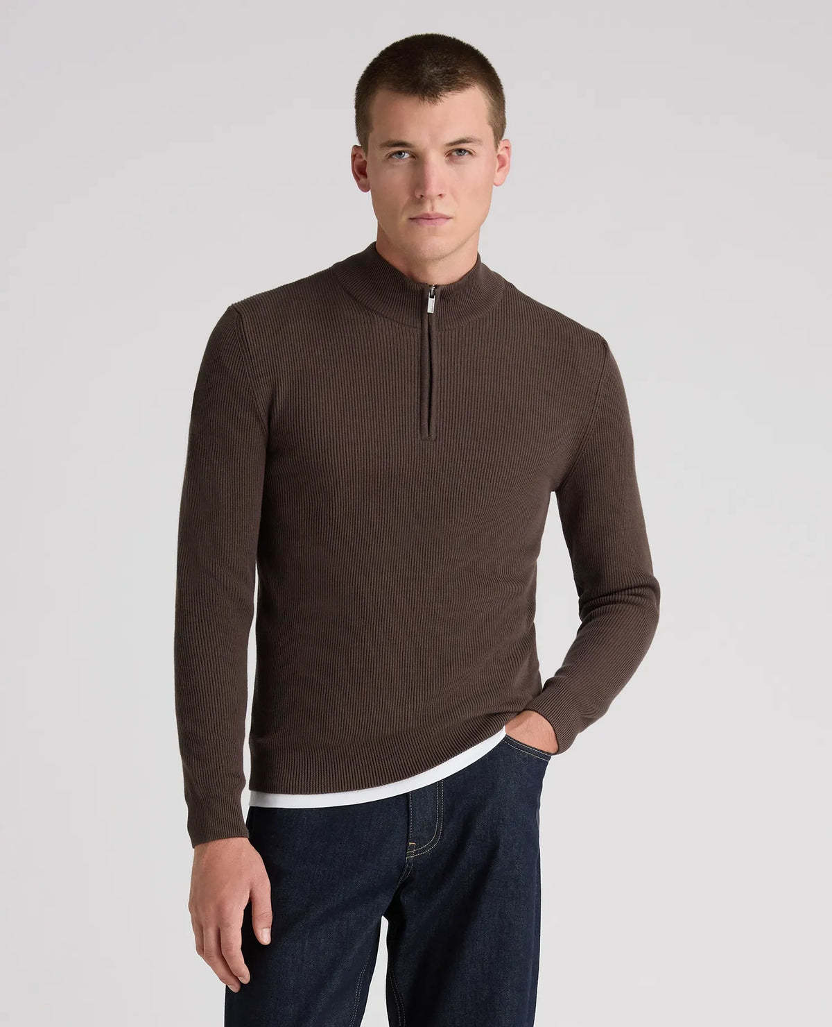 A man wearing a dark brown ribbed quarter-zip sweater with a mock neck, paired with dark blue jeans, photographed against a neutral light gray background.