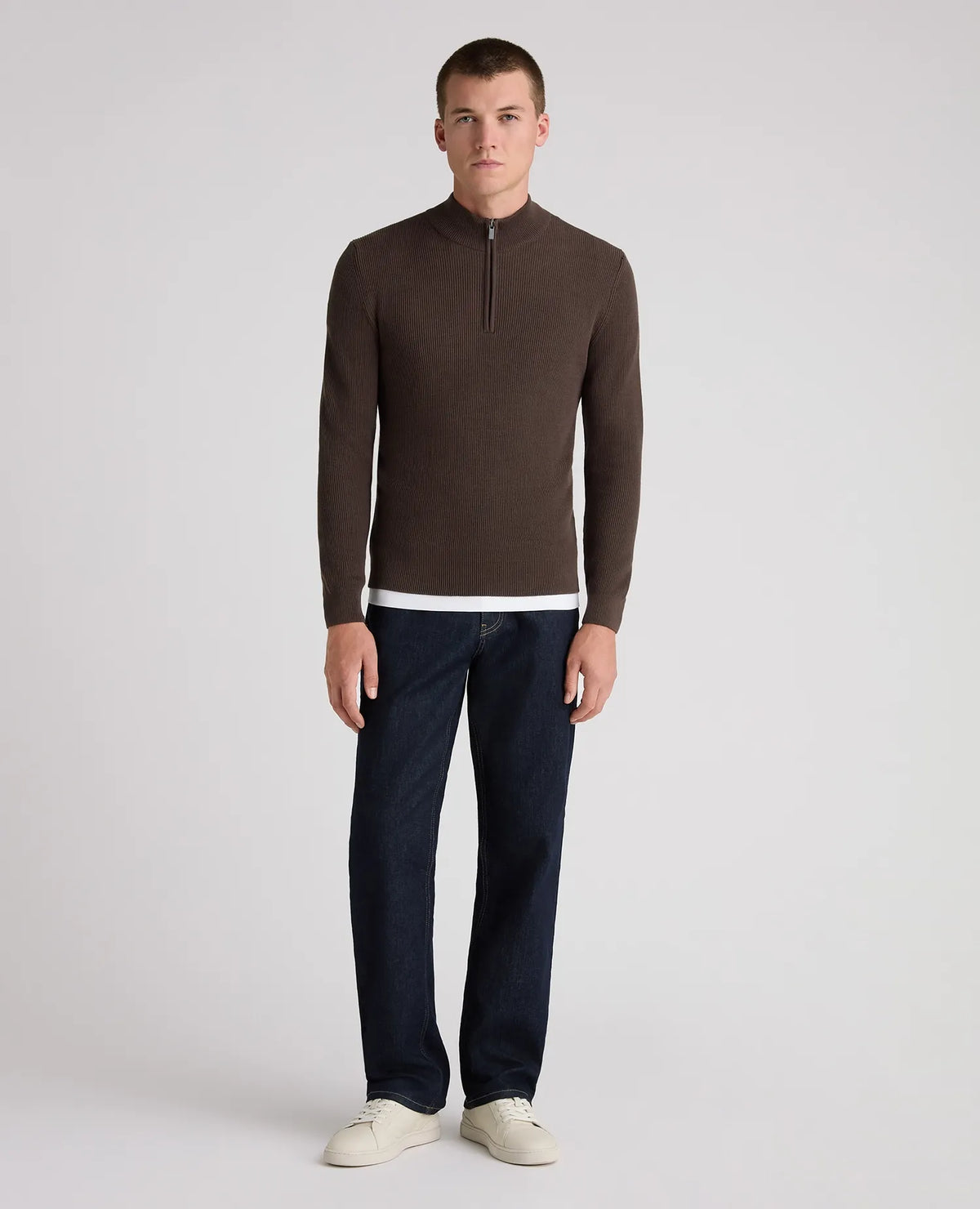 A man modeling a dark brown quarter-zip knit sweater with a white hem detail, paired with dark blue jeans and off-white sneakers, against a light gray studio background.