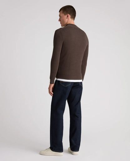 A man is shown from the back wearing a dark brown long-sleeve knit sweater, dark blue jeans, and off-white sneakers against a plain light gray studio background.