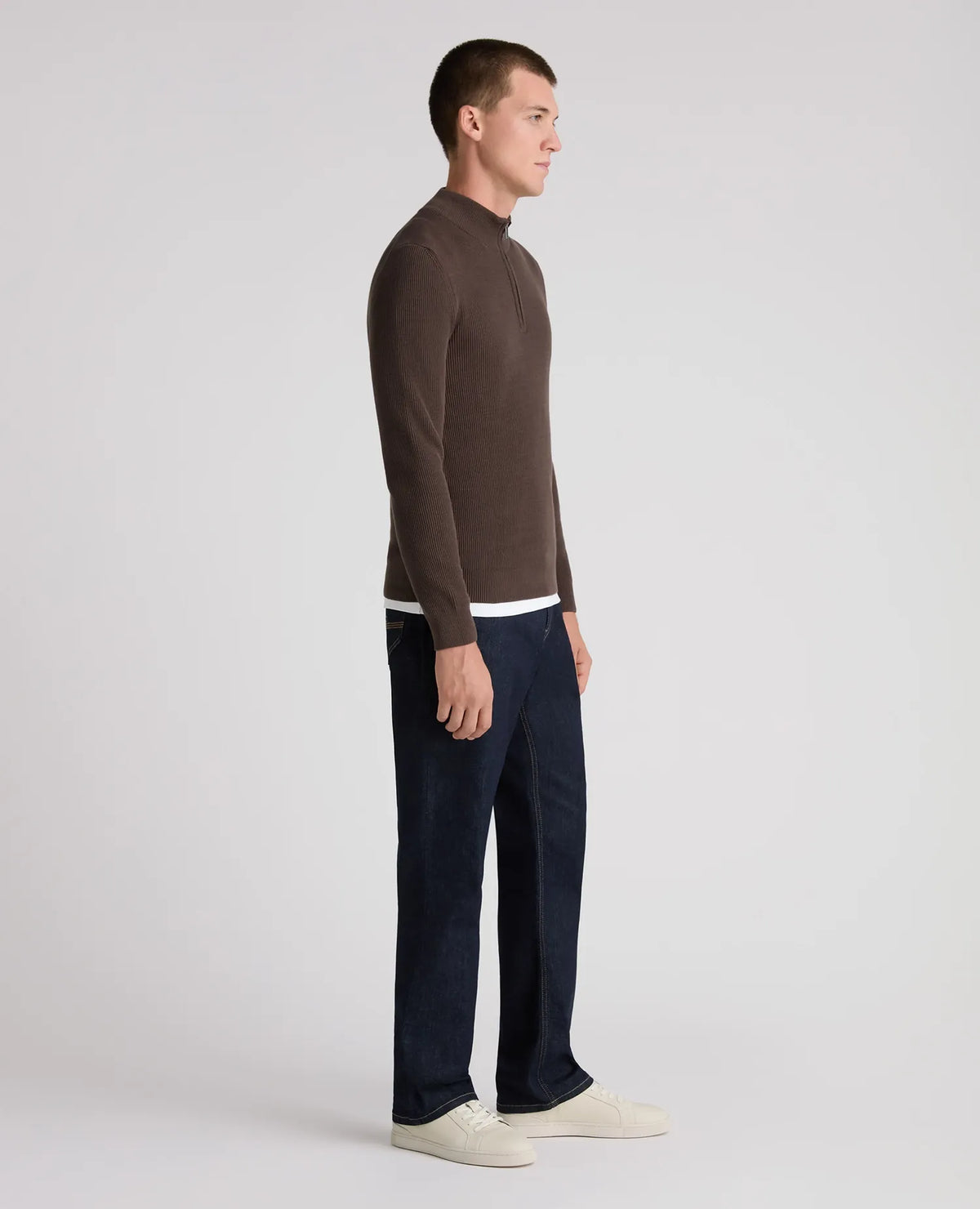 A man in profile wearing a brown mock-neck long-sleeve sweater, dark navy blue jeans, and off-white sneakers, standing against a light gray studio background.