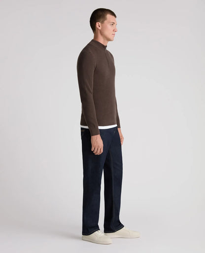 A man in profile wearing a brown mock-neck long-sleeve sweater, dark navy blue jeans, and off-white sneakers, standing against a light gray studio background.