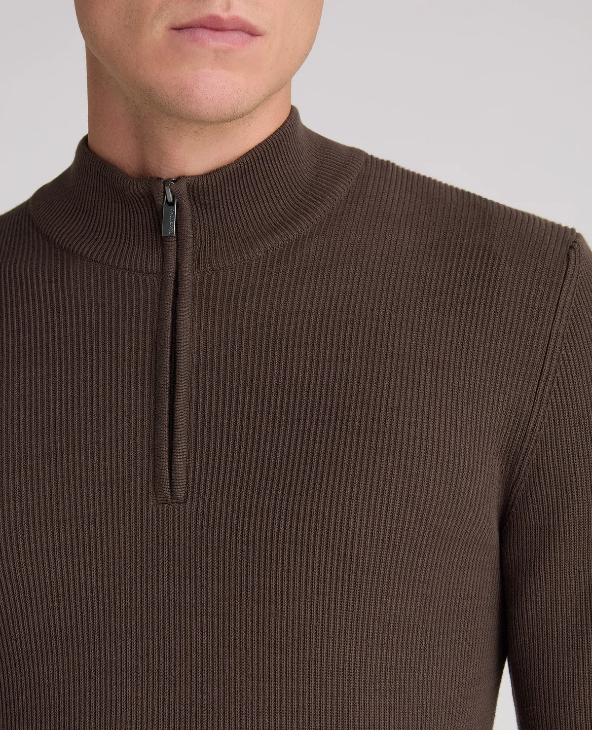 Close-up of a man wearing a ribbed brown half-zip mock neck sweater against a light gray background.