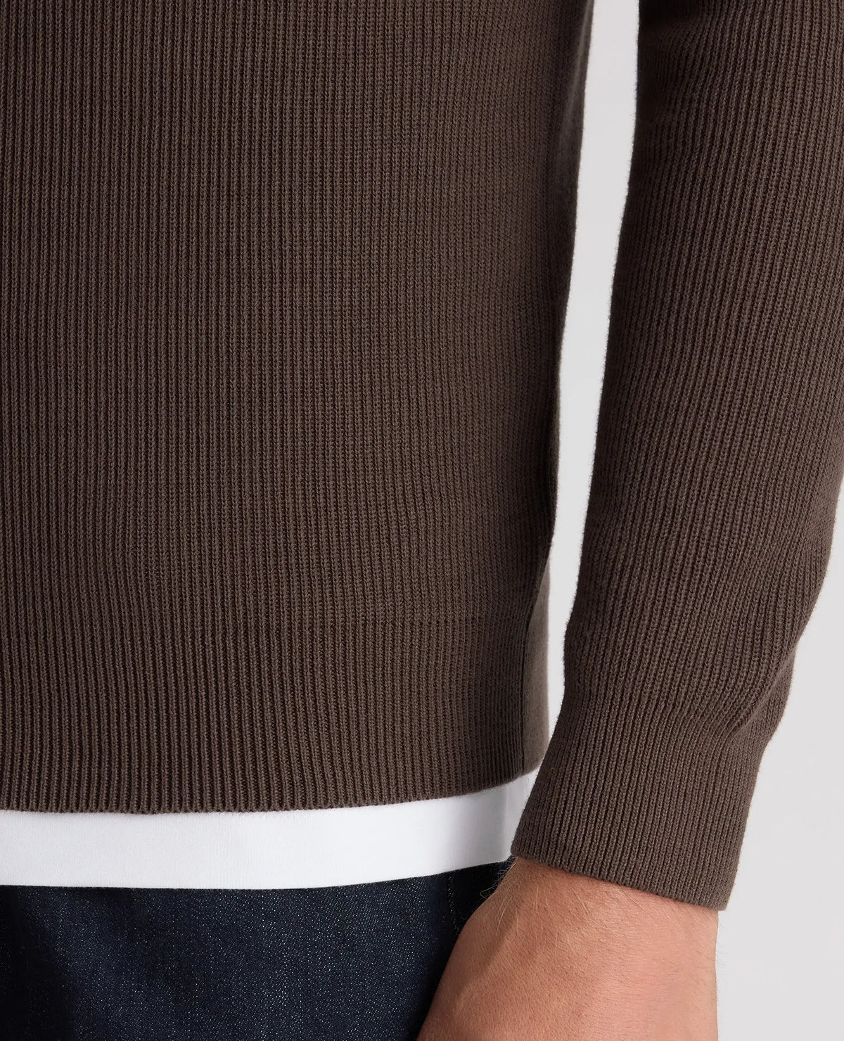 Close-up of a ribbed knit sweater in dark brown, showing the sleeve cuff, side seam, and a white undershirt hem peeking out above dark blue jeans.