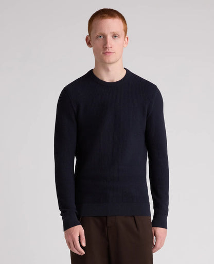 A man with red hair wearing a dark navy ribbed knit crewneck sweater and dark brown trousers, standing against a light gray studio background.