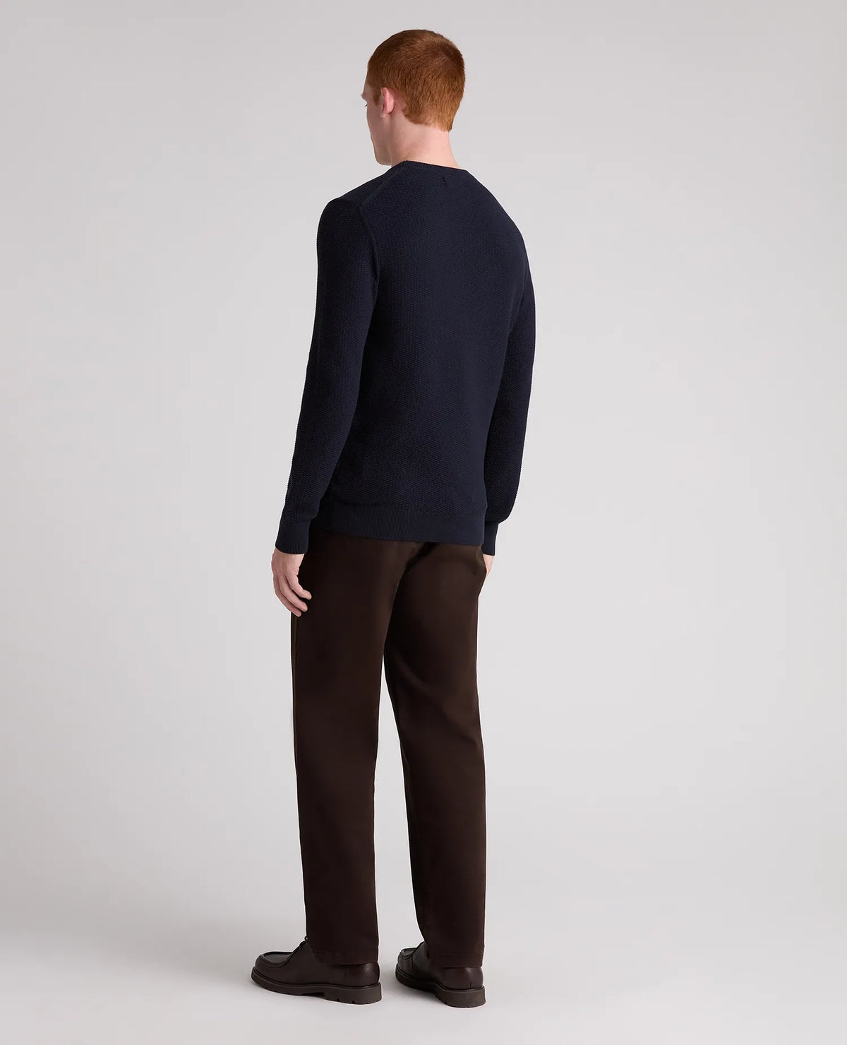 A man with red hair is shown from the back wearing a dark navy blue long-sleeve crewneck sweater and dark brown trousers, standing against a light gray studio background.