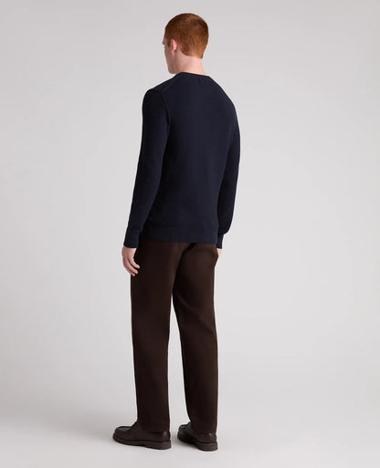 A man with red hair is shown from the back wearing a dark navy blue long-sleeve crewneck sweater and dark brown trousers, standing against a light gray studio background.