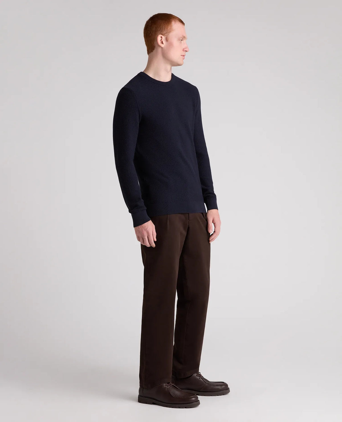 A man in profile wearing a dark navy long-sleeve crewneck sweater, dark brown trousers, and dark brown leather shoes, standing against a light gray studio background.