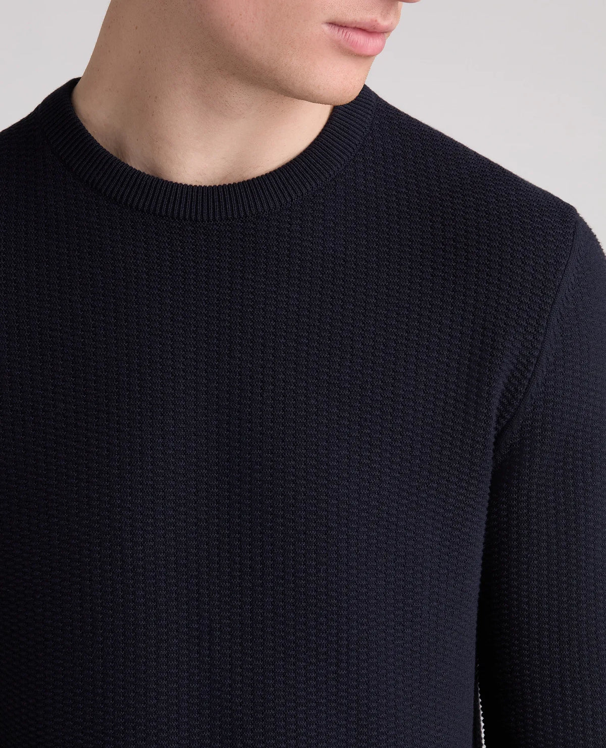 Close-up of a person wearing a dark navy textured knit crewneck sweater against a light gray background.