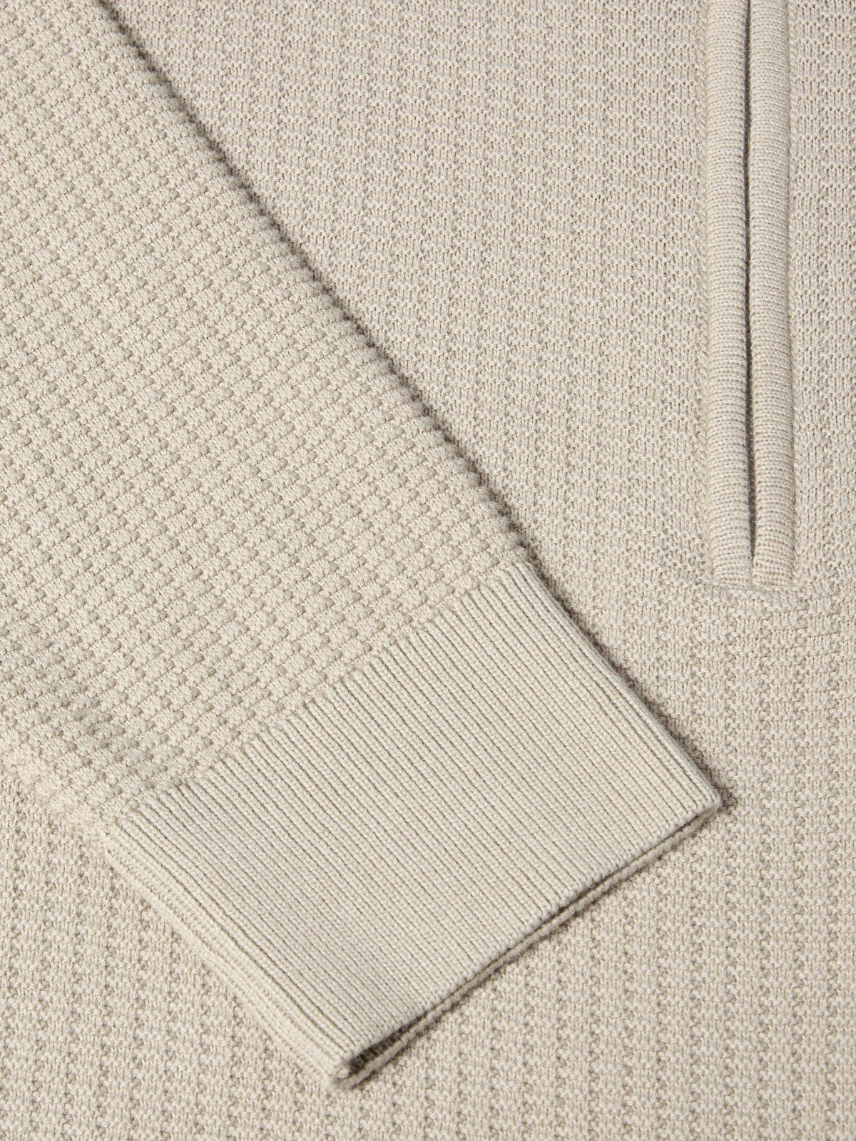 Close-up of a cream-colored knit sweater showing the waffle texture, a ribbed cuff, and part of a vertical ribbed placket.